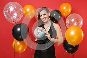 Smiling young woman in little black dress celebrating and holding world globe on bright red background air balloons