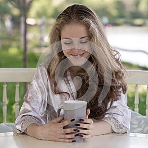 Smiling young woman with long curly hair dressed in silk peignoir is drinking a coffee in a white pavilion in the garden.