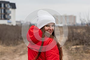Smiling young woman with long hair in white knitted hat and red jacket with hood looking over her shoulder on the street