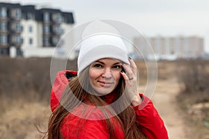 Smiling young woman with long hair in white knitted hat and red jacket with hood looking over her shoulder on the street