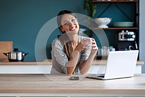 Smiling young woman looking at camera while holding a cup of coffee and working with laptop in the kitchen at home