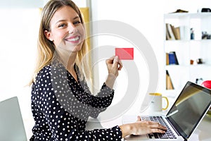 Smiling young woman looking at camera while holding red credit card for shopping online with computer at home