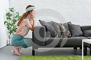 smiling young woman looking at her tabby cat while he sitting on couch