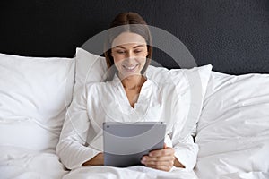 Smiling young woman lying in comfortable bed, using digital tablet.