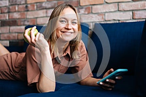 Smiling young woman lying on sofa using smartphone at home.