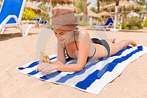 Smiling young woman is lying on striped towel on the sand at the beach and applying sun cream on her hand