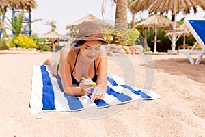 Smiling young woman is lying on striped towel on the sand at the beach and applying sun cream on her hand