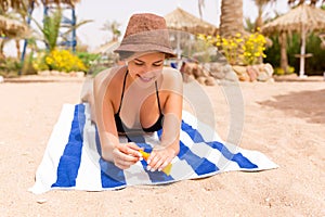 Smiling young woman is lying on striped towel on the sand at the beach and applying sun cream on her hand