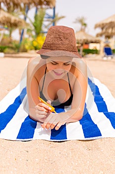 Smiling young woman is lying on striped towel on the sand at the beach and applying sun cream on her hand