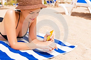 Smiling young woman is lying on striped towel on the sand at the beach and applying sun cream on her hand