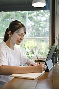 Smiling young woman making note on notebook and using computer tablet at coffee shop.