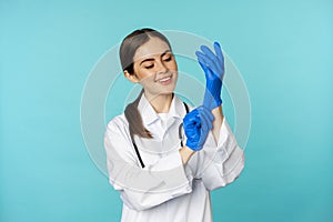 Smiling young woman, medical worker wearing gloves for patient checkup, standing in hospital clinic uniform over blue