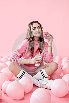Smiling young woman with milkshake and donut in hands sitting on floor with many pink air balloons