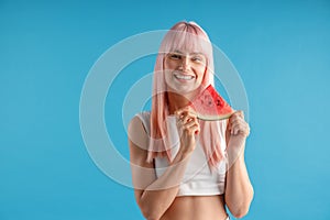 Smiling young woman with natural long pink dyed hair holding a slice of watermelon, posing isolated over blue studio