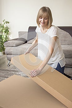 Smiling young woman opens cardboard boxes in the living room. Sofa and flower in the background.