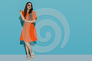 Smiling young woman in orange cocktail dress and high heels sandals standing against a blue sunny wall