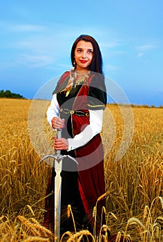 Smiling Young woman with ornamental dress and sword in hand standing on a wheat field with sunset. Natural background..