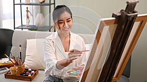 Smiling young woman painting picture on canvas with oil paints in bright cozy living room. Art, creative hobby and