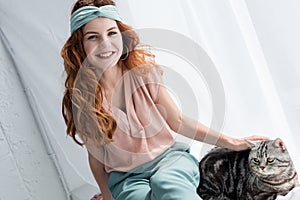 smiling young woman petting adorable tabby cat while sitting