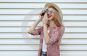 Smiling young woman photographer with vintage film camera wearing summer round straw hat, checkered shirt on white wall