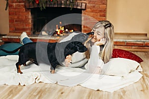 A smiling young woman is playing with a dog, sitting on the floor near the fireplace, on Christmas Eve. Christmas mood