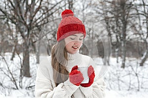 Smiling young woman in red knitted hat and mittens holds cup of hot tea outdoors. Warm drinks in mug in winter walk