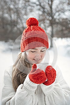 Smiling young woman in red knitted mittens holds cup of hot drink outdoors. Warm drinks in winter