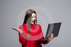 Smiling young woman in red sweater holding laptop computer and pointing at copyspace while looking at the camera over gray