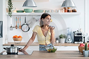 Smiling young woman sending an audio with her mobile phone while eating a salad in the kitchen at home