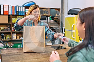 Smiling young woman shop assistant delivering paper bag with purchased items to female customer