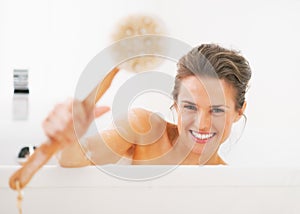 Smiling young woman showing body brush in bathtub