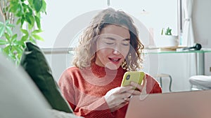 Smiling young woman sitting on couch using cell phone doing online shopping.