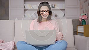 Smiling asian woman sitting on sofa using laptop computer looking at screen typing message, mixed race lady chatting on