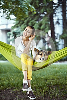 Smiling young woman sitting in green hammock with cute dog Welsh Corgi in a park outdoors. Beautiful happy female in