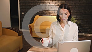 Smiling young woman sitting at home with a cup of coffee, using laptop, has video call chatting, happy woman chatting on