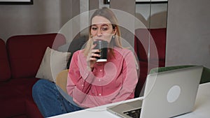 Smiling young woman sitting at home with a cup of coffee, using laptop, has video chatting, happy woman chatting on