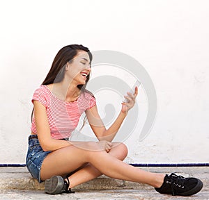 Smiling young woman sitting on sidewalk reading text on cell phone