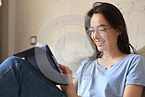 Smiling young woman sitting on sofa with digital tablet and chating with friends.