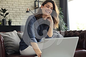 Smiling young woman sitting on sofa with laptop computer and chating with friends