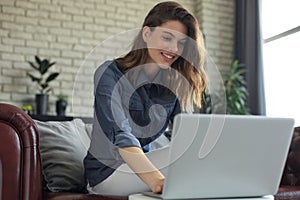 Smiling young woman sitting on sofa with laptop computer and chating with friends