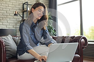 Smiling young woman sitting on sofa with laptop computer and chating with friends