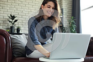 Smiling young woman sitting on sofa with laptop computer and chating with friends