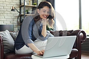 Smiling young woman sitting on sofa with laptop computer and chating with friends