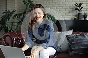 Smiling young woman sitting on sofa with laptop computer and chating with friends