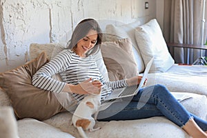 Smiling young woman sitting on sofa with laptop computer and chating with friends, little dog playing near.