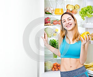 Smiling young woman staing with fruits and vegetables near the fridge full of healthy food.