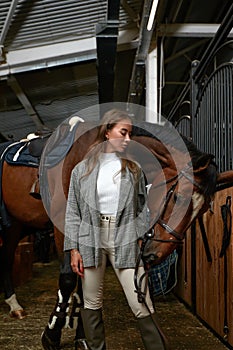 Smiling young woman standing inside a stable holding a saddle while preparing her chestnut horse for a ride