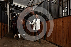 Smiling young woman standing inside a stable holding a saddle while preparing her chestnut horse for a ride