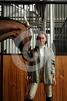 Smiling young woman standing inside a stable holding a saddle while preparing her chestnut horse for a ride