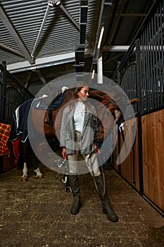 Smiling young woman standing inside a stable holding a saddle while preparing her chestnut horse for a ride